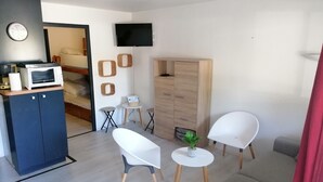 Een moderne woonkamer met een bank, twee witte stoelen, een bijzettafeltje met een plant, een aan de muur bevestigde televisie, en een kitchenette met een magnetron en broodrooster.
