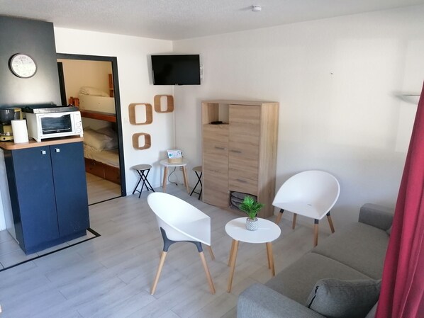 Un salon moderne comprenant un canapé, deux fauteuils blancs, une petite table avec une plante, un téléviseur fixé au mur et un coin cuisine équipé d’un micro-ondes et d’un grille-pain.