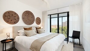 A modern hotel room with a large bed, a side table, and a chair. There are three woven wall art pieces above the bed. - Fox on Macleay is a Two Bedroom Apartment (Elizabeth Bay)