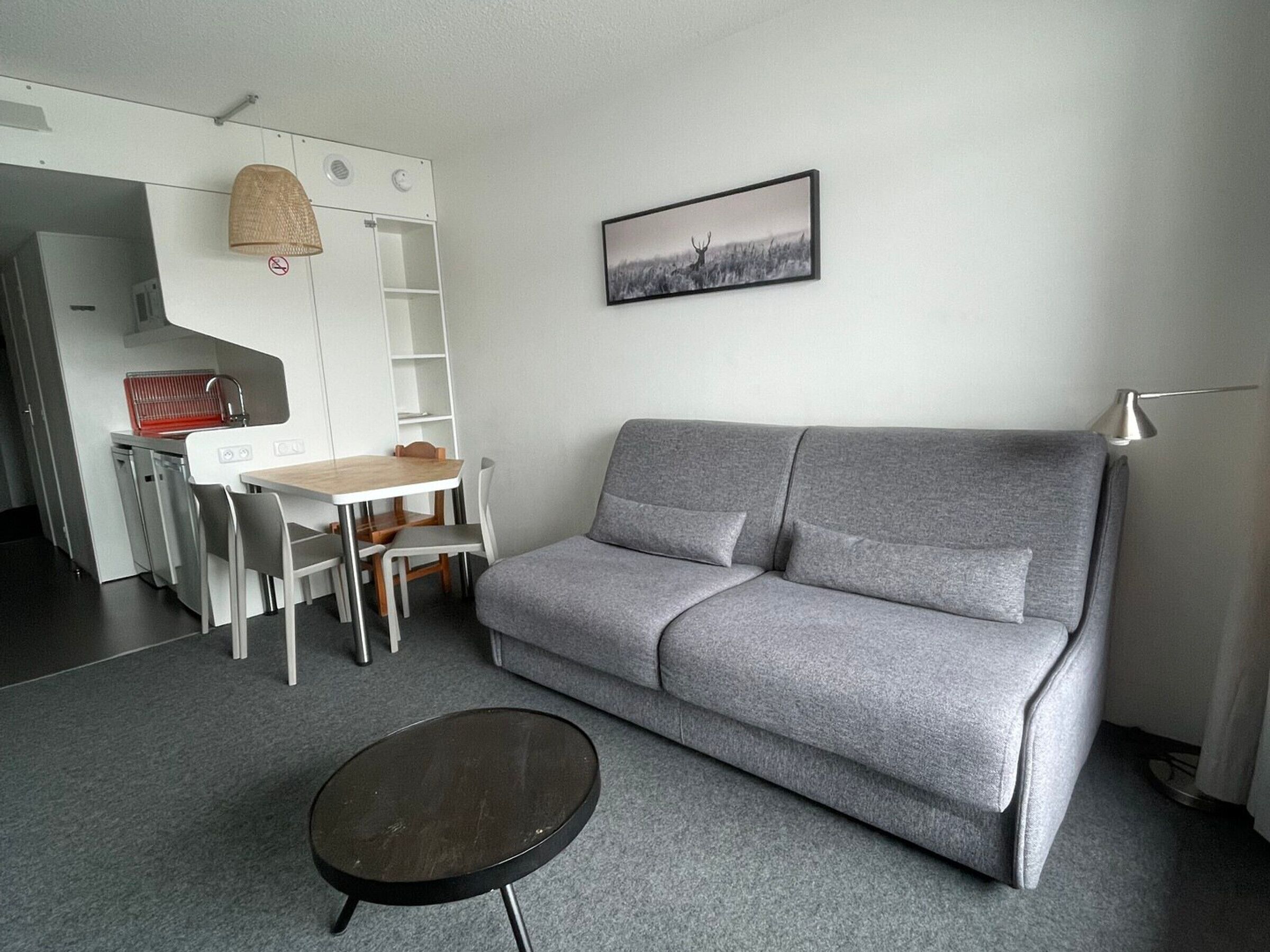 A modern living room with a grey sofa, a small round table, a dining area with a white table and chairs, and a kitchen area with a red dish rack.