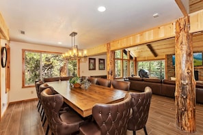 A spacious dining area with a wooden table and leather chairs, large windows with a view of trees, and a stone fireplace. - Gold Rush Lodge: Exceptional Location in Big Bear's Eagle Mountain Estates! Luxury 5 En Suite Bedrooms! EV Charger! Pet Friendly! (Big Bear Lake)