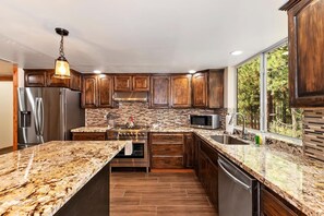A modern kitchen with granite countertops, stainless steel appliances, wooden cabinets, and a large window providing a view of the outdoors.