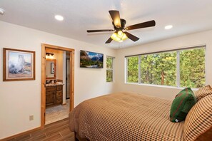 A bedroom with a bed, a ceiling fan, a window with a view of trees, and a door leading to a bathroom.