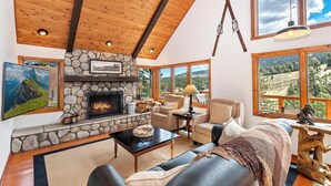 Un salón acogedor con chimenea de piedra, techo de madera y grandes ventanas con vistas panorámicas.