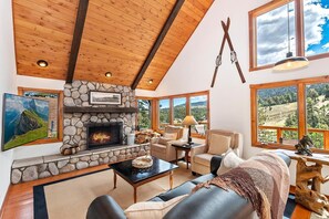 A cozy living room with a stone fireplace, wooden ceiling beams, and large windows offering a view of the outdoors.