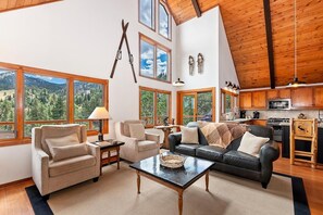 A spacious living area with a large window, a fireplace, and a kitchen in the background. - Aspen Escape by Destination Big Bear: Incredible Views and Game Table. Close to Bear Mountain and the Zoo! EV Charger! (Big Bear Lake CA)