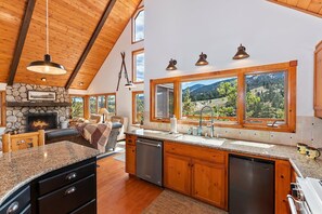 A spacious kitchen with a stone fireplace, wooden cabinets, and large windows offering a scenic view. - Aspen Escape by Destination Big Bear: Incredible Views and Game Table. Close to Bear Mountain and the Zoo! EV Charger! (Big Bear Lake CA)