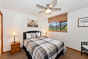 A bedroom with a bed, two bedside lamps, a ceiling fan, a window with a view, and a framed picture on the wall. - Aspen Escape by Destination Big Bear: Incredible Views and Game Table. Close to Bear Mountain and the Zoo! EV Charger! (Big Bear Lake CA)