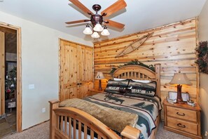Un dormitorio con cabecera de madera, cama con manta estampada, mesita de noche con lámpara y ventilador de techo.