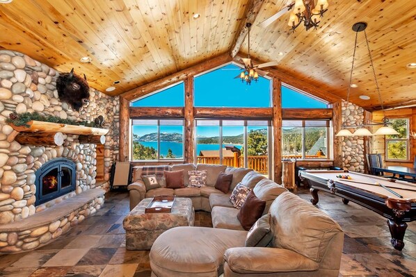 A spacious room with a stone fireplace, a pool table, and a large window offering a view of the outdoors.