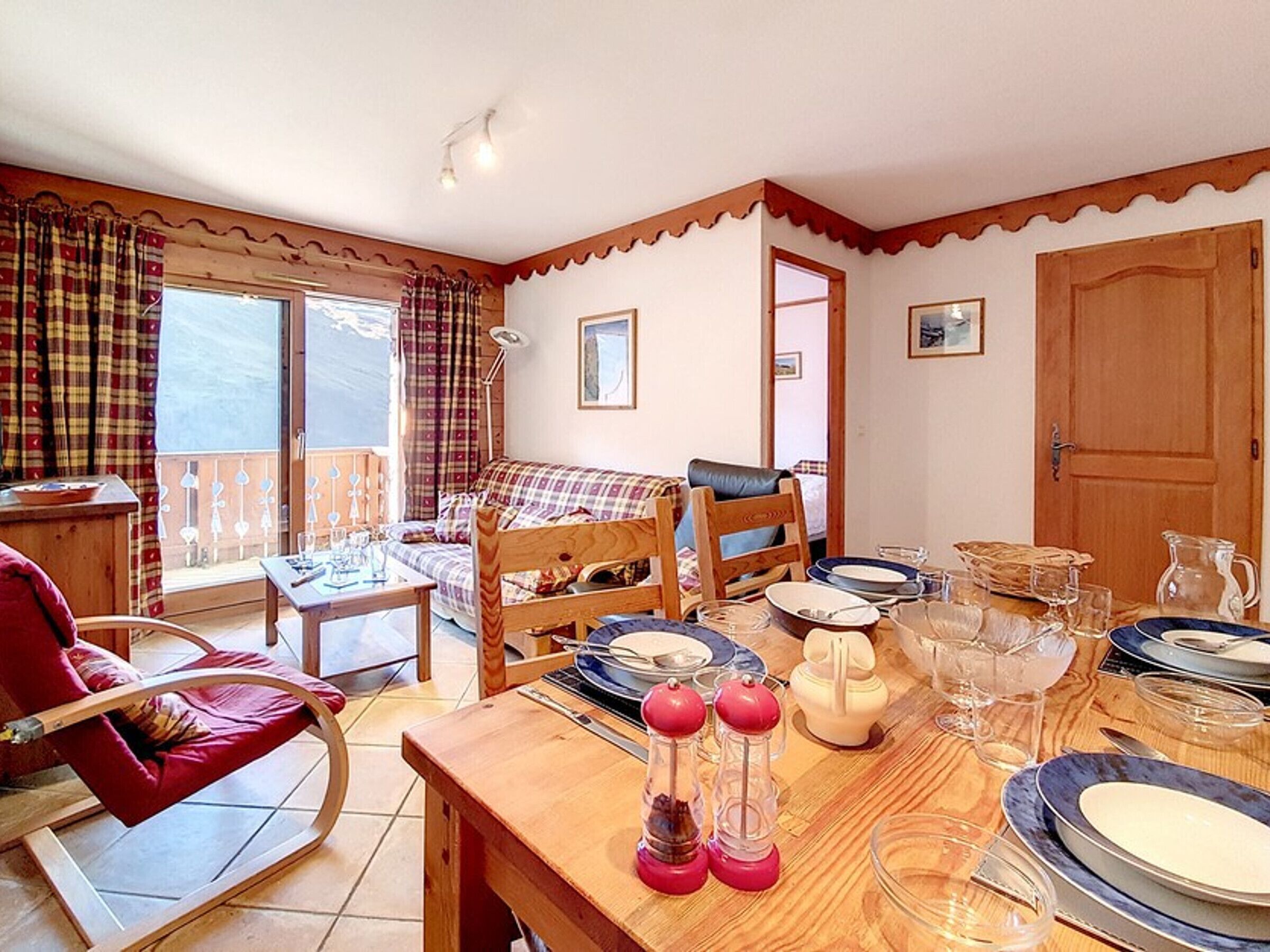 A dining area with a wooden table set for four, a red chair, a sofa, and a balcony with curtains.