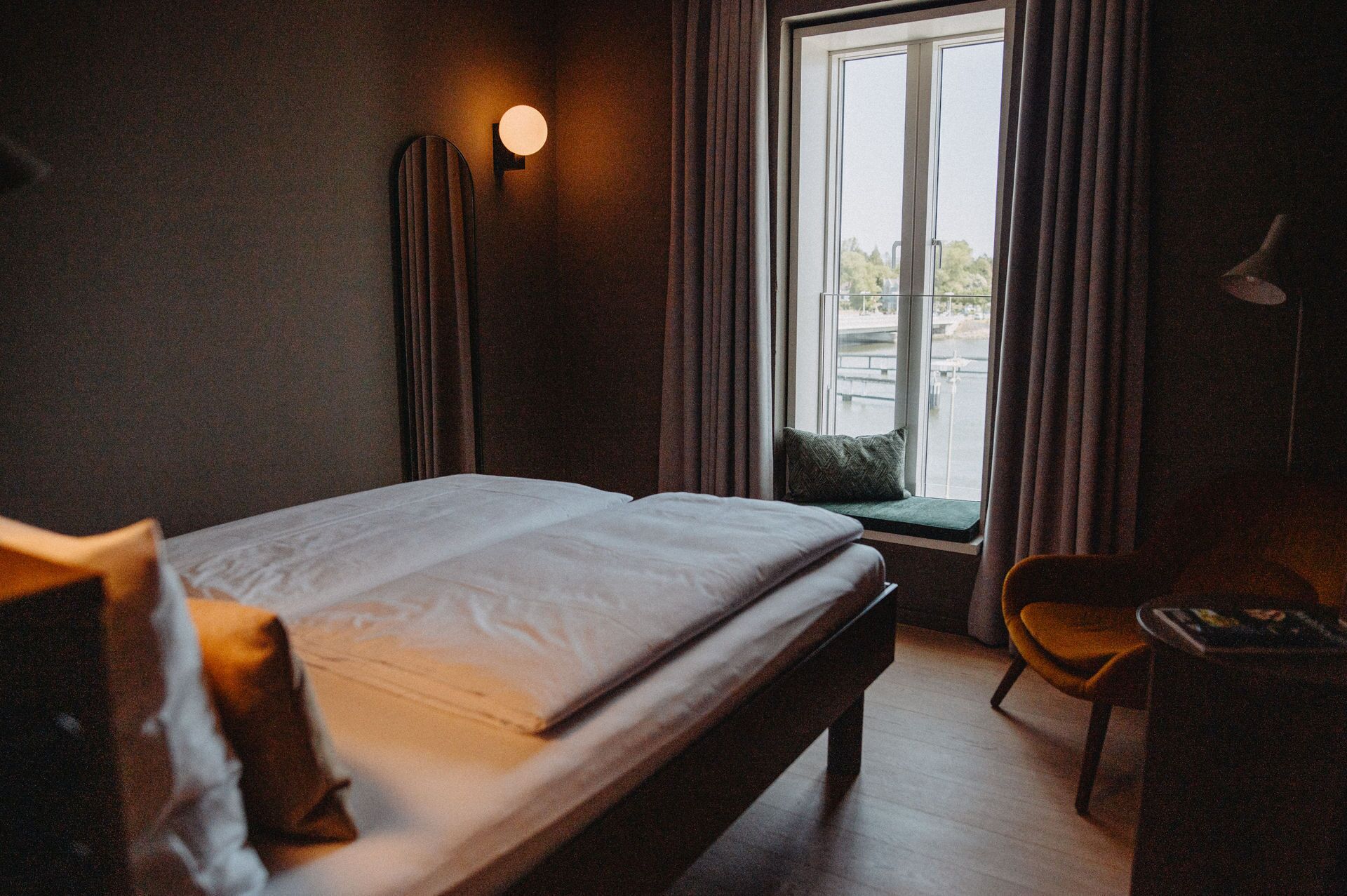A hotel room with a bed, a desk, a chair, a window with curtains, and a wall-mounted light.