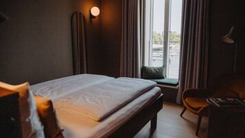 A hotel room with a bed, a desk, a chair, a window with curtains, and a wall-mounted light.
