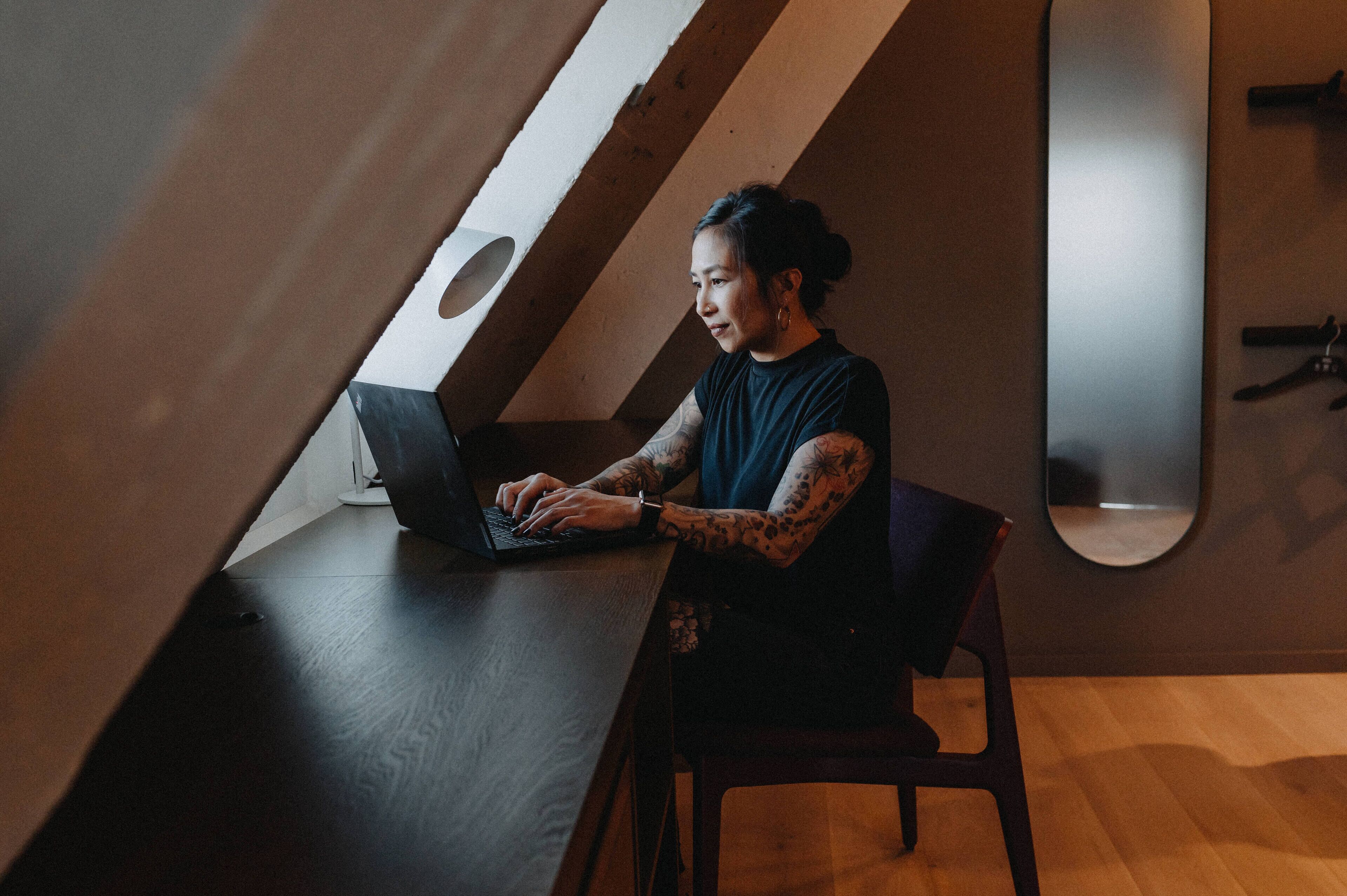 Eine Person sitzt an einem Schreibtisch mit einem Laptop in einem Raum mit schrägen Decken und einem großen Spiegel an der Wand.