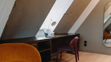 A modern loft bedroom with a sloped ceiling, a wooden desk with a laptop and lamp, a red chair, and a large mirror on the wall.