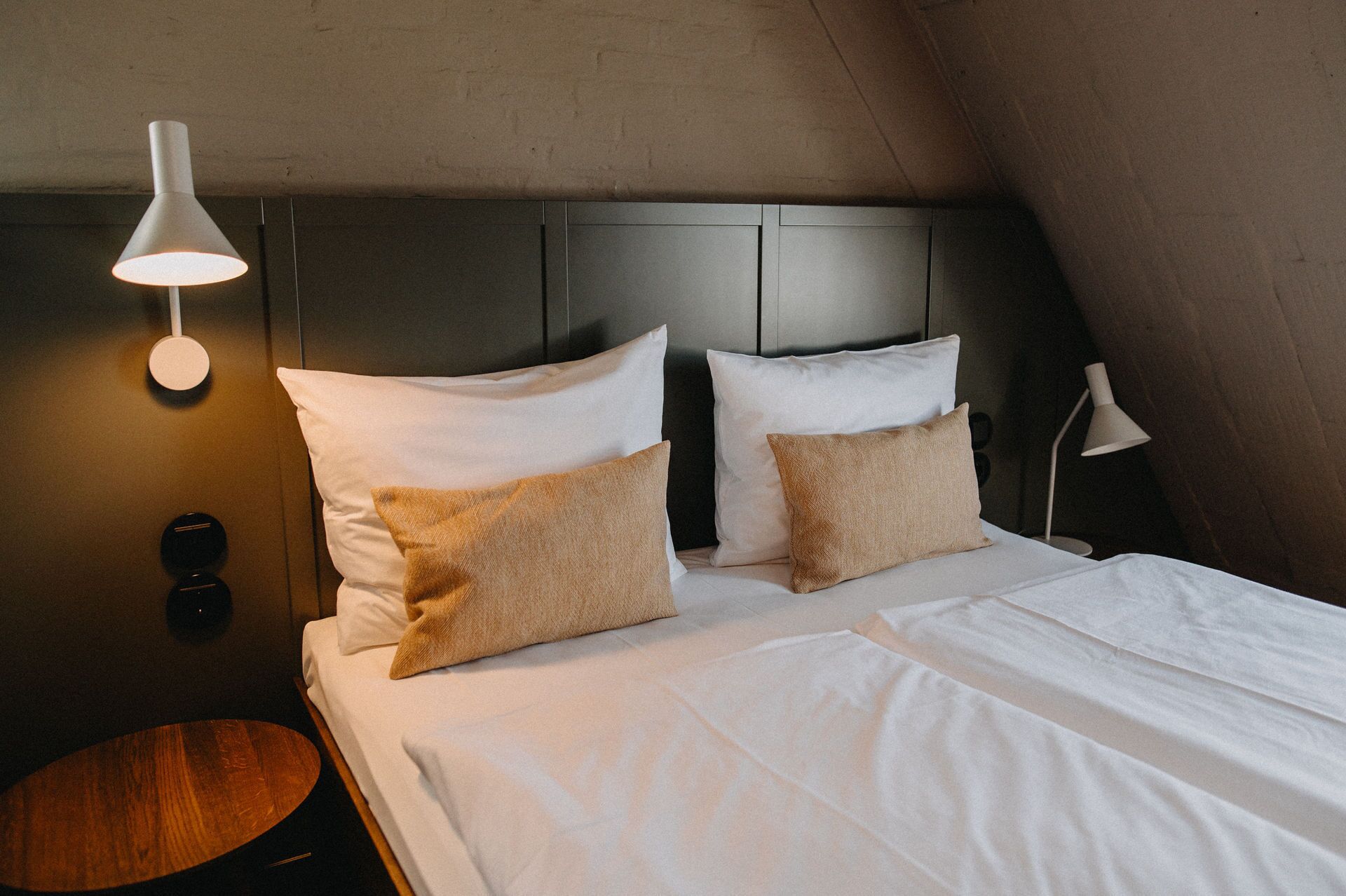 Un lit bien fait, avec du linge de lit blanc et deux oreillers beige, une table ronde en bois et une lampe moderne sur le côté gauche.