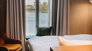 A hotel room with a bed, a chair, a window with curtains, and a view of the water. - Südspeicher (Kappeln)