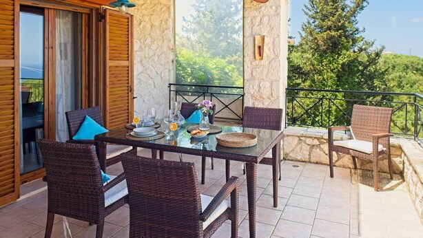 An outdoor dining area with wicker furniture, a glass table, and a stone wall backdrop. - Villa Petalouda Stunning Villa in Tragaki Zante (Zakynthos)