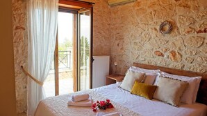 A bedroom with a stone wall, a bed with white linens, a window with curtains, and an air conditioning unit. - Villa Petalouda Stunning Villa in Tragaki Zante (Zakynthos)