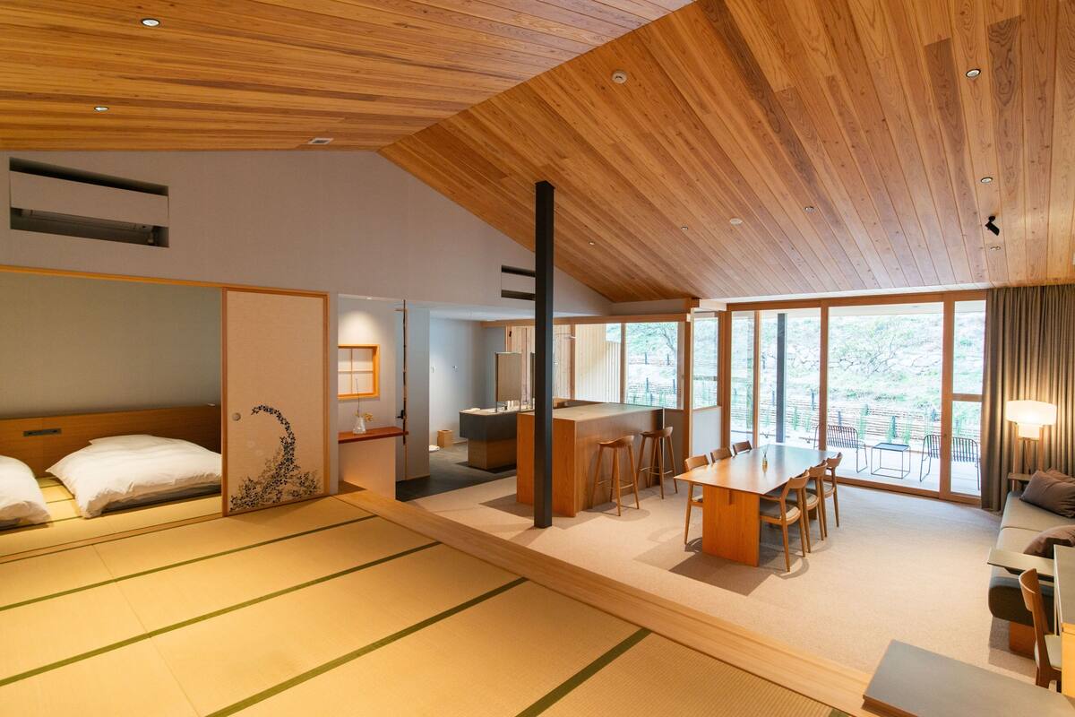 A modern hotel room with tatami mats, a wooden ceiling, a dining area, and large windows.