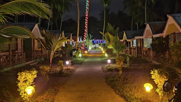 A pathway leading to a row of thatched-roof cottages, surrounded by palm trees and illuminated by ground lights. - Honey Moon Beach Resort (Port Blair)