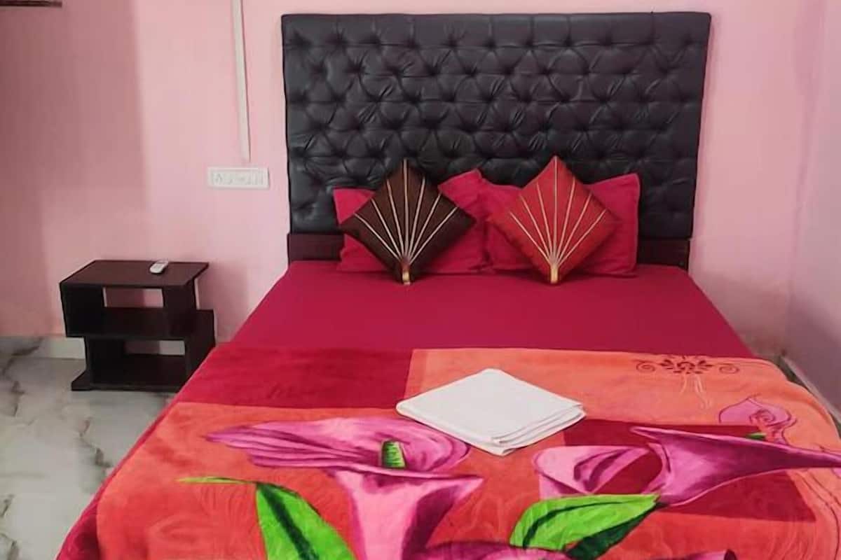Una cama con cabecera capitonada, colcha roja con estampado floral y una mesita de noche con control remoto.