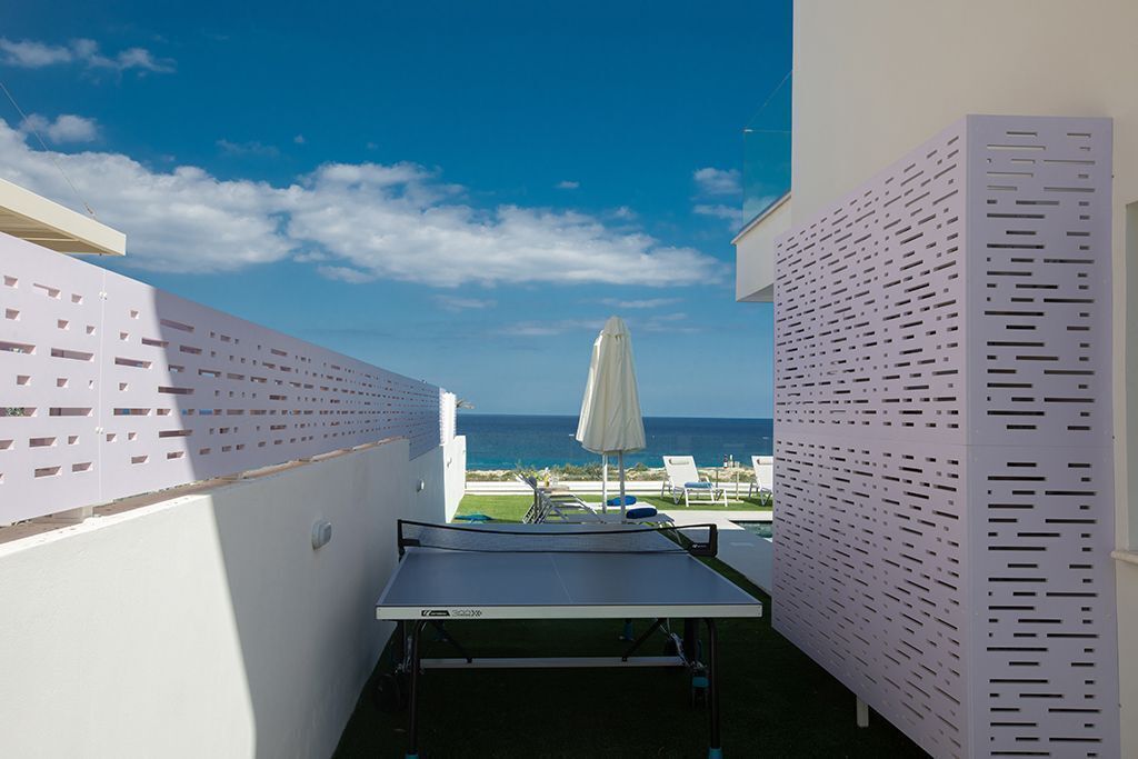 Ein Billardtisch in einer sonnendurchfluteten Außenbereich mit Blick aufs Meer, weißen Wänden und einer durchbrochenen Sichtschutzwand.