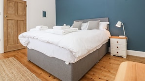 A neatly made bed with white linens and pillows, a bedside table with a lamp, and a wooden floor. - Angorfa (Pwllheli)