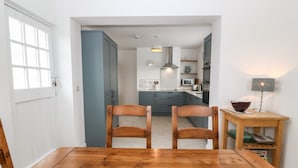 A dining area with a wooden table and chairs, a kitchen in the background, and a lamp on a side table. - Angorfa (Pwllheli)