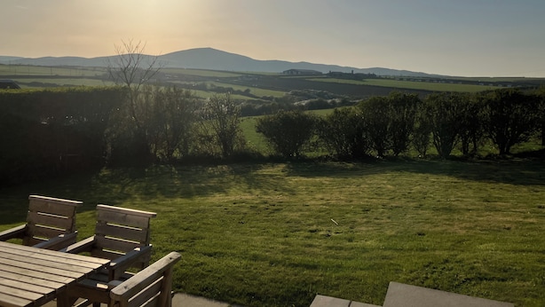 A grassy outdoor area with a wooden table and chairs, a clear view of distant hills, and a bright sun in the sky. - Angorfa (Pwllheli)