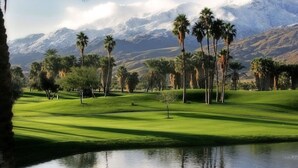 A golf course with a water body in the foreground, palm trees, and snow-capped mountains in the background.