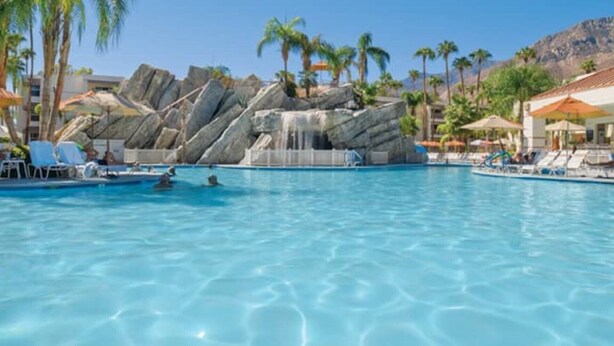 A swimming pool with clear blue water, surrounded by lounge chairs and umbrellas, with a rocky waterfall feature in the background.