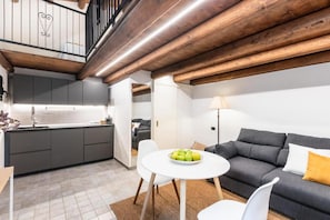 A modern kitchen with grey cabinets, a white countertop, and a wooden ceiling. There is a dining area with a white table and chairs, and a grey sofa with a yellow pillow. - Modern and Spacious Apartment in Noto, Sicily (Noto)