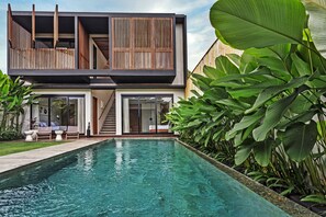 A modern two-story house with a swimming pool, wooden deck, and lush greenery.