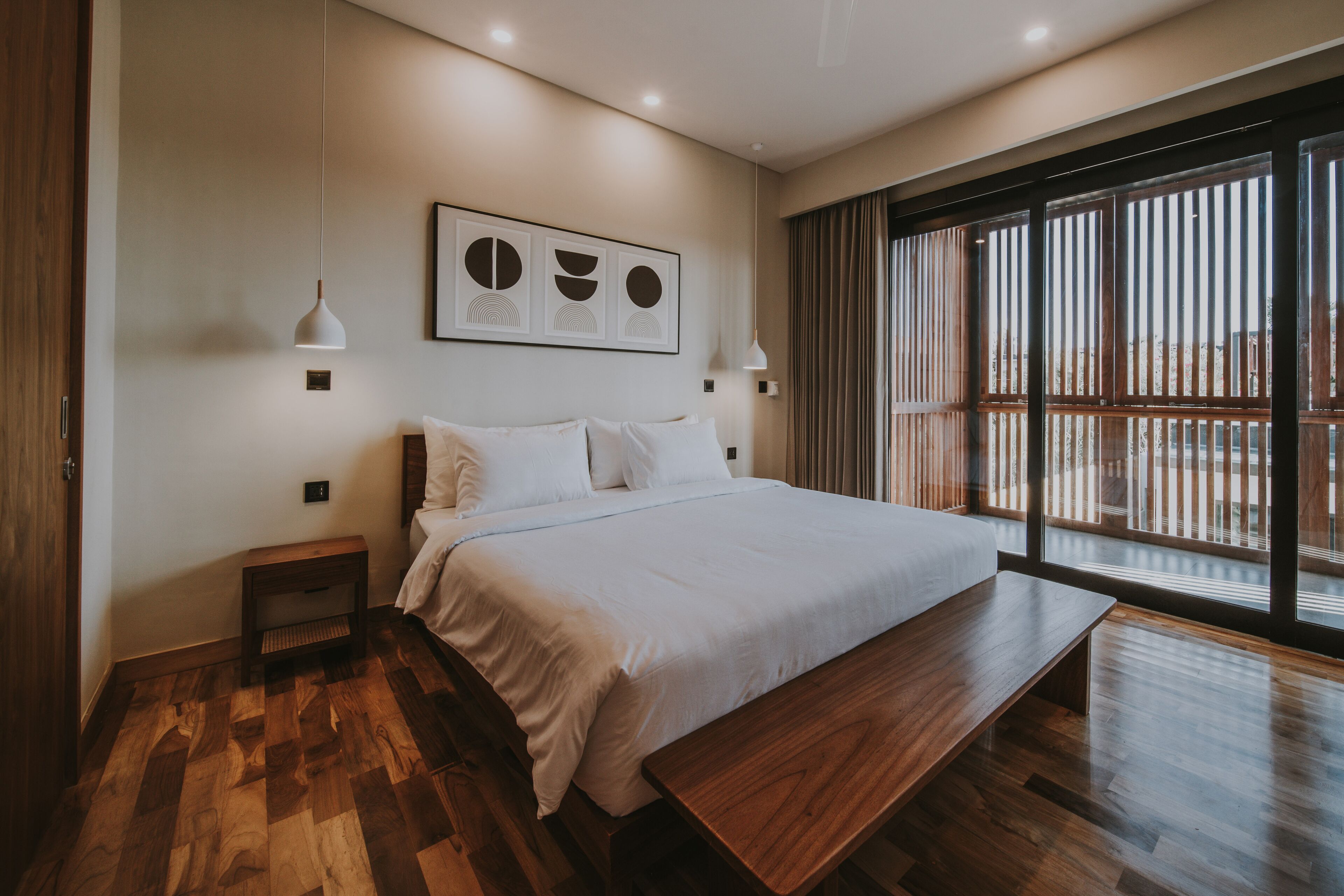 A modern bedroom with a large bed, wooden flooring, a bench, and a large window with blinds.