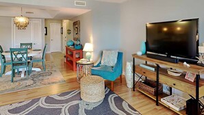 A living room with a TV, blue sofa, and a dining area with teal chairs. - Inlet Point 15B, Beautifully Furnished Condo, Near Beach & Creek (Pawleys Island)