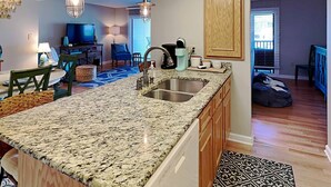 A modern kitchen with granite countertops, stainless steel sink, and wooden cabinets. There is a dining area with a blue chair and a TV in the background. - Inlet Point 15B, Beautifully Furnished Condo, Near Beach & Creek (Pawleys Island)