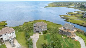 A coastal property with two main buildings, a white house and a grey house with a balcony, surrounded by grassy areas and a large body of water. - Windscape Soundfront in Frisco Incredible Views 400ft to Beach Access! (Frisco)