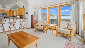 A spacious living room with a wooden coffee table, two sofas, and a dining area with wooden chairs. Large windows offer a view of the outdoors. - Windscape Soundfront in Frisco Incredible Views 400ft to Beach Access! (Frisco)