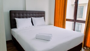 Uma cama bem arrumada com lençóis brancos e uma toalha enrolada, uma cabeceira escura e uma janela com cortinas laranja.