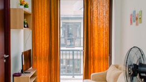Uma sala de estar moderna com um sofá, uma mesinha, uma televisão e um ventilador. Há uma janela com cortinas, uma porta e uma estante com objetos decorativos.
