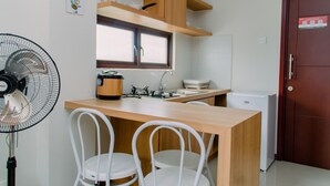Cozinha compacta com bancada de madeira, fogão a gás, pia, pequena geladeira e ventilador. Há duas cadeiras brancas e uma janela sobre a pia.