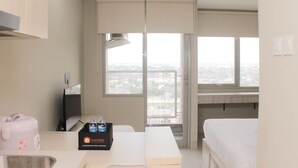 A hotel room with a bed, a desk with a computer, a window with a city view, and a minibar. - Nice Studio at LRT City Jatibening Apartment (Bekasi)