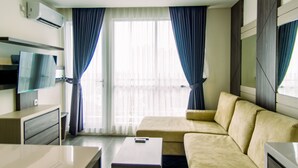 A modern hotel room with a beige sofa, a flat-screen TV mounted on the wall, a small table, and a window with blue curtains. - Comfort And Best Choice 2Br At Bintaro Icon Apartment (South Tangerang)