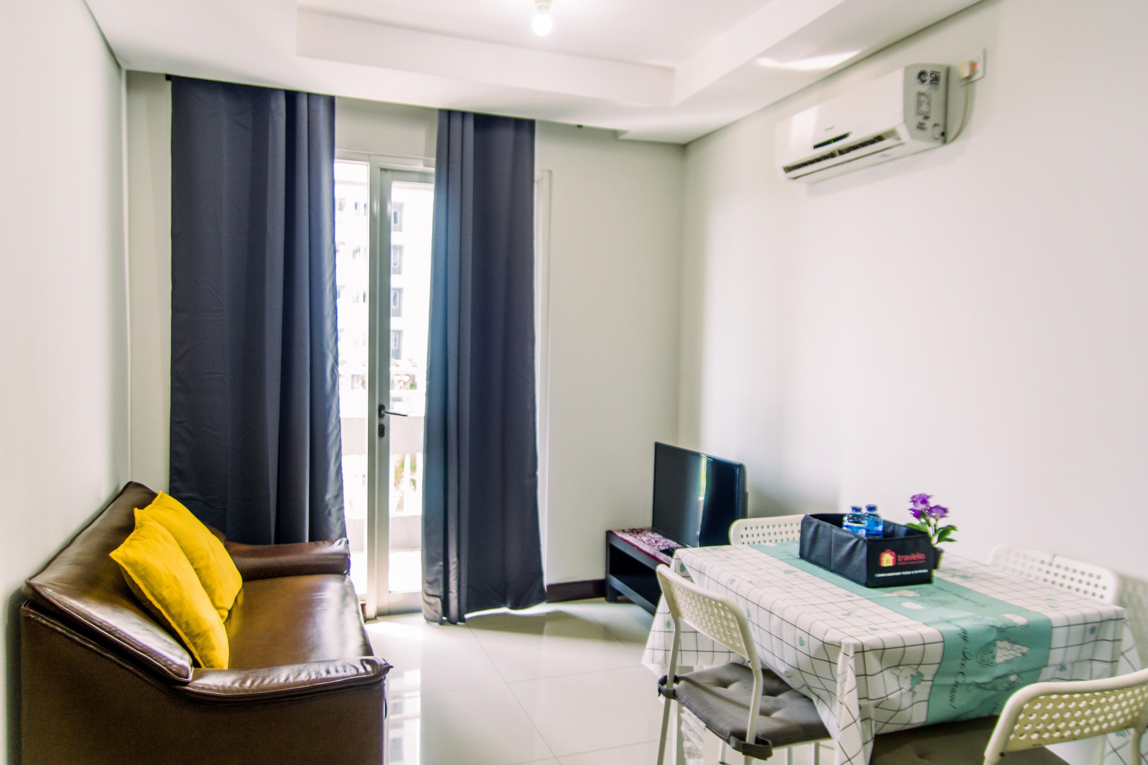 A room with a brown leather sofa, a dining table with a white tablecloth, a TV, and an air conditioning unit on the wall.