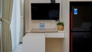 A modern room with a flat-screen TV mounted on a wall, a black refrigerator with a blue sticker, a small plant, and two water bottles on a white counter.