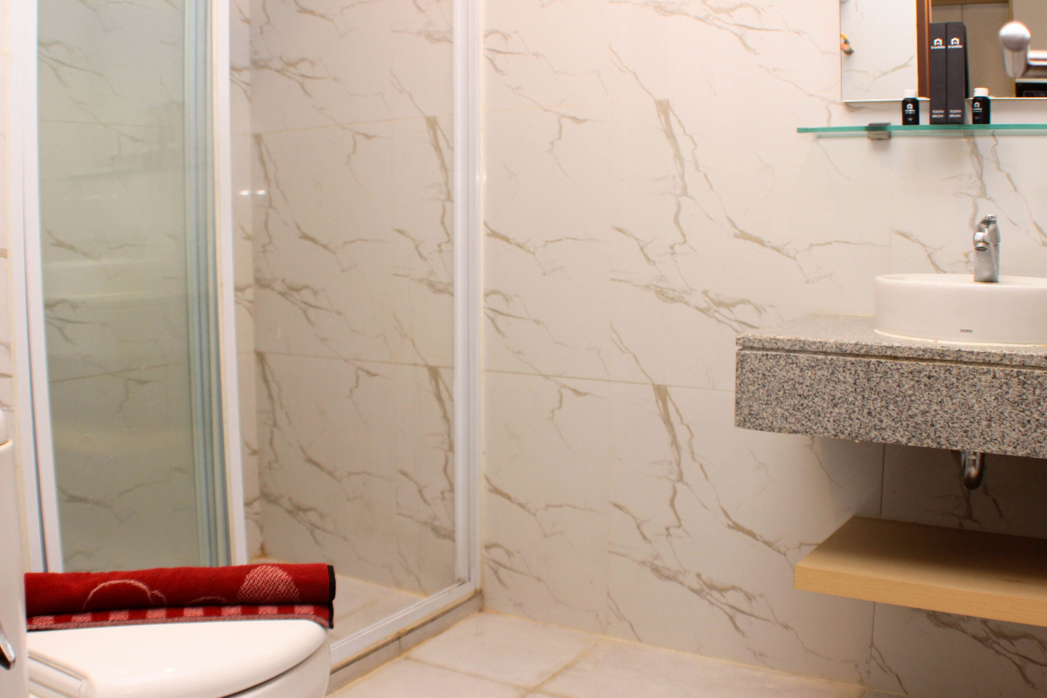 A bathroom with a glass shower enclosure, a white toilet, a marble wall, a granite sink, and a shelf with toiletries.