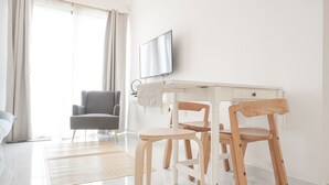 A modern, well-lit room with a dining area featuring a white table and wooden chairs, a grey sofa, and a television mounted on the wall. - Homey And High Floor 2Br At Sky House Bsd Apartment (Pagedangan)