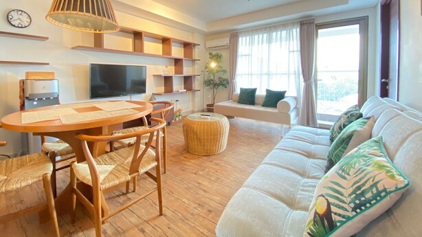 A modern living room with a wooden dining table, wicker chairs, a sofa with cushions, a TV, and built-in shelves. - Spacious 2Br Apartment Tamansari Tera Residence (Bandung)