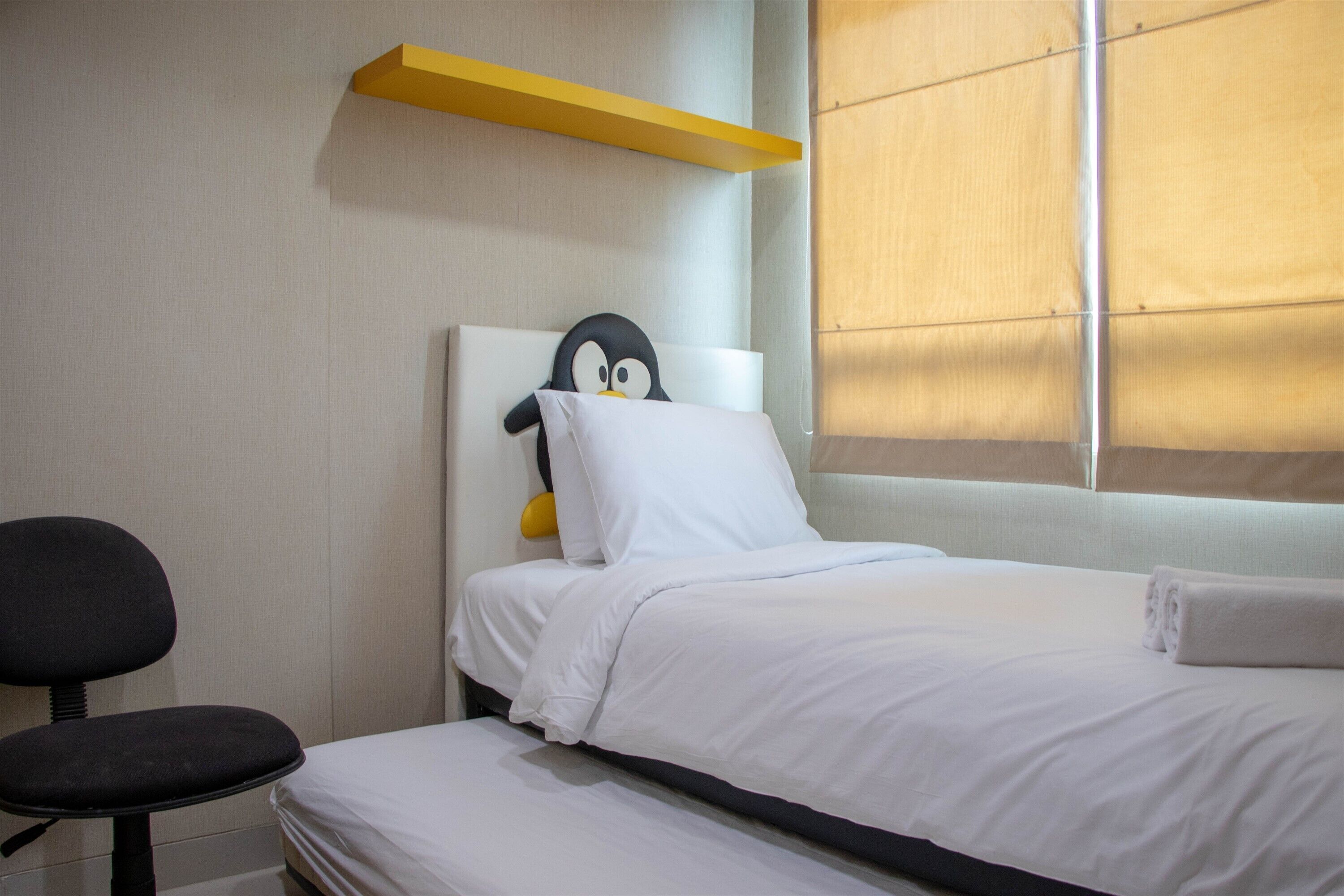 A hotel room with a bed, a chair, a pillow with a penguin, a yellow shelf, and a window with blinds.